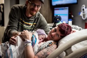 Danielle Fetty-Lovell, a white-presenting person with short dark red hair, snuggles her newborn baby while reclined in a hospital bed, with her husband nearby